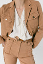Person wearing a veste coton beige and matching high-waisted pants with frayed seams, paired with a white ruffled blouse. The outfit features gold button details and a black belt. The person’s face is not visible.