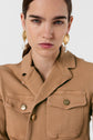 A woman with straight brown hair wears gold earrings with pearls and a tan veste saharienne buttoned jacket, standing against a plain white background, looking directly at the camera with a neutral expression.