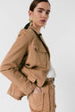 Person wearing a veste saharienne—tan jacket and matching pants with fringe details—a white top, and large gold earrings, posing with one hand in their pocket against a plain white background.