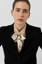 Person with light skin and light brown hair, wearing a veste noire de tailleur, beige shirt, and patterned silk scarf tied at the neck, poses against a plain white background for a look that defines dressing chic.