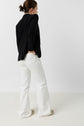 Person standing with back to the camera, wearing a veste noire de tailleur, white pants, and beige shoes, with one hand in a pocket and hair tied back. The background is plain white, creating an effortlessly dressing chic look.