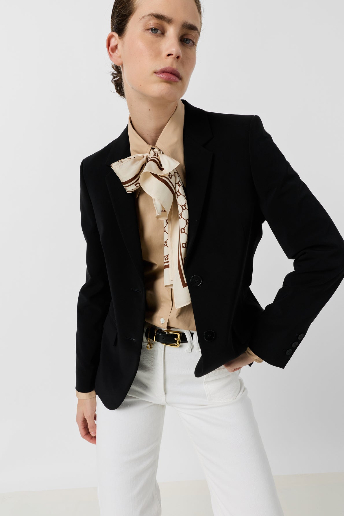 Person wearing a veste de tailleur noire, beige blouse with a patterned neck scarf, white pants, and black belt, standing against a plain white background with one hand on their hip and a confident expression—an effortlessly dressing chic look.