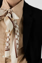 A close-up of a person wearing a beige shirt, a patterned silk scarf tied in a bow, and a veste noire de tailleur. The scarf features geometric and letter designs in brown on a cream background, creating an effortlessly dressing chic look.