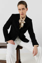 A person in a veste noire, beige blouse with a patterned scarf, and white pants sits on a wooden bench, leaning slightly forward with a serious expression against a plain background—a perfect example of dressing chic.