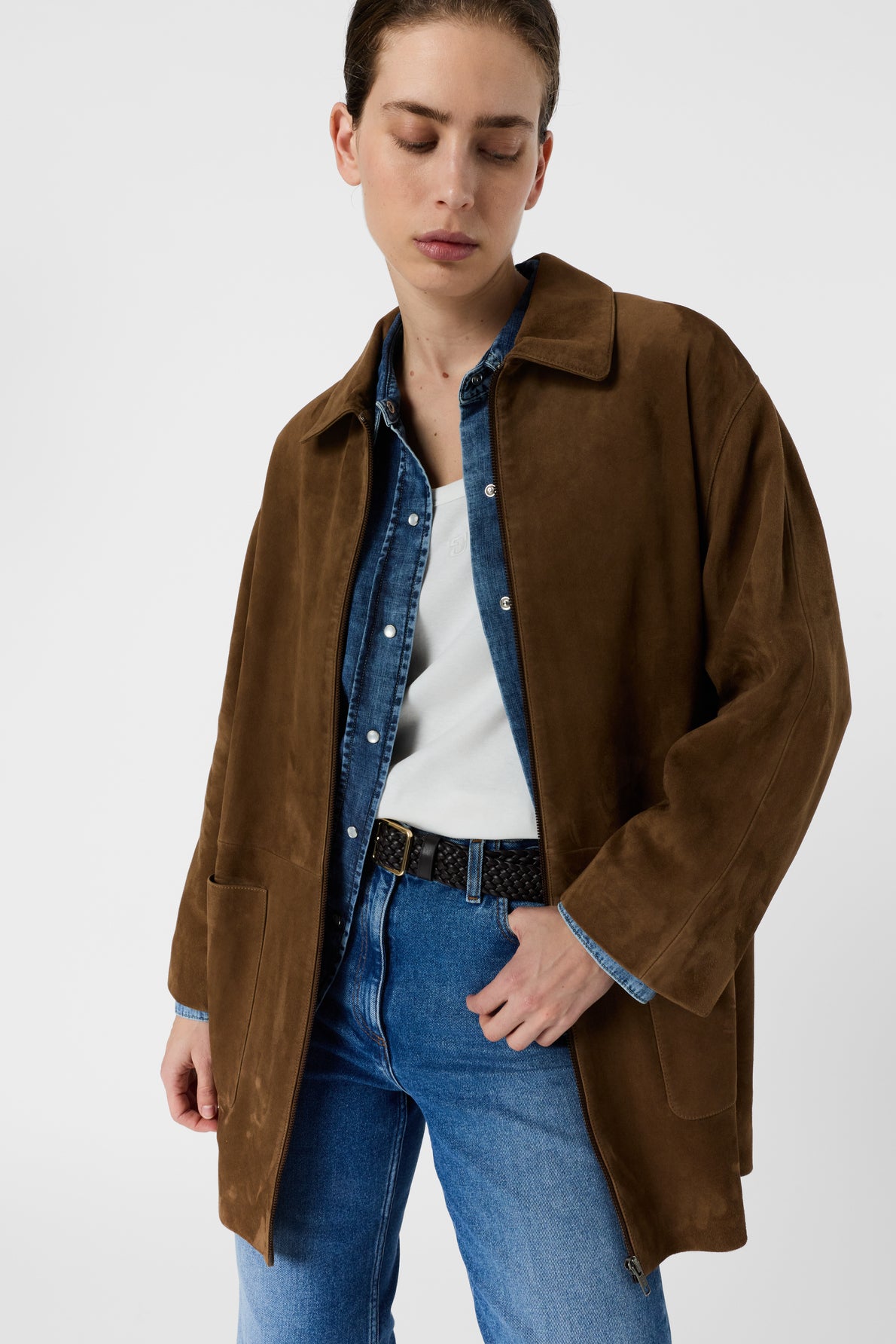 A person wearing a brown suede jacket over a denim shirt, white top, and blue jeans stands against a plain white background with eyes closed and head slightly tilted down.