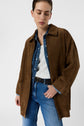 A person wearing a brown suede jacket over a denim shirt, white top, and blue jeans stands against a plain white background with eyes closed and head slightly tilted down.
