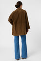 A person with hair in a low bun wears a brown suede jacket and blue jeans, standing facing away from the camera against a plain white background.
