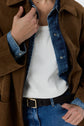 A person wears a brown jacket over a blue denim shirt and white t-shirt, paired with blue jeans and a black braided belt. Their hand adjusts the jacket collar.