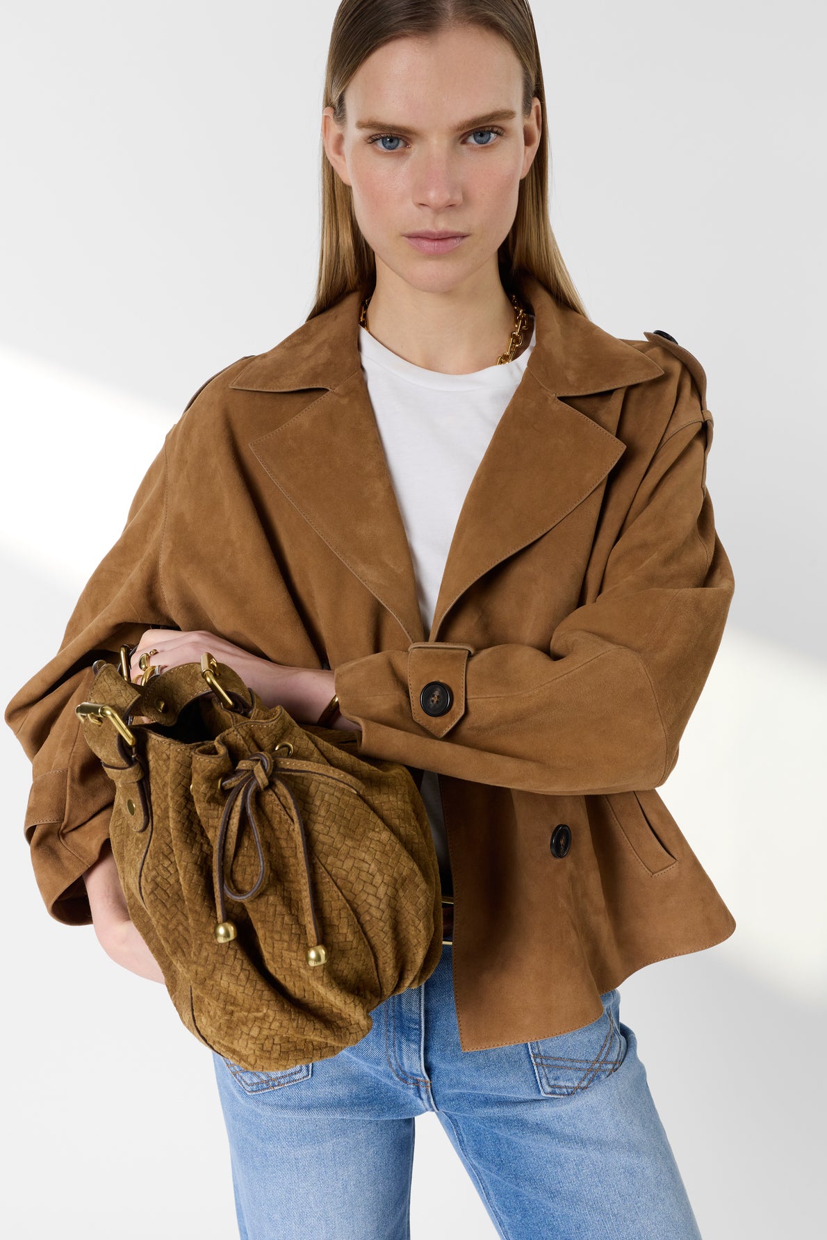 A woman wearing a brown suede jacket, white t-shirt, and blue jeans holds a textured brown handbag. She has straight, light brown hair and a neutral expression, standing against a plain, light background.