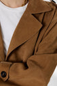 Close-up of a person wearing a tan suede trench coat over a white shirt. A gold chain necklace is partly visible, and the coat features a buttoned cuff. The photo is cropped at the shoulders.