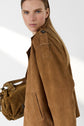 A person with long blonde hair wears a brown suede jacket and holds a matching brown textured bag, looking at the camera with a neutral expression. The background is plain and brightly lit.