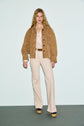 Woman with long blonde hair wears a veste en cuir velours camel, light striped blouse, cream flared pants, a brown belt, and platform shoes. She stands against a plain background, looking at the camera.