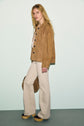 A woman with long blonde hair wears a veste en cuir velours, cream wide-leg pants, and platform shoes, standing against a pale background and holding a matching tan bag.