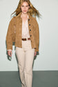 A person with long blonde hair wears a veste en cuir velours camel, a striped shirt, beige high-waisted pants, and a brown belt, carrying a tan shoulder bag while walking against a light background.