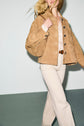 A woman with long blonde hair wears a veste en cuir velours, paired with light beige pants and a tucked-in light shirt. She is walking, her face partially out of frame. The background is plain and light-colored.