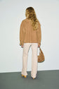 A woman with long, wavy blonde hair stands facing away, wearing a veste camel over cream-colored pants, tan platform shoes, and holding a tan suede bag. The background is plain and minimalistic.