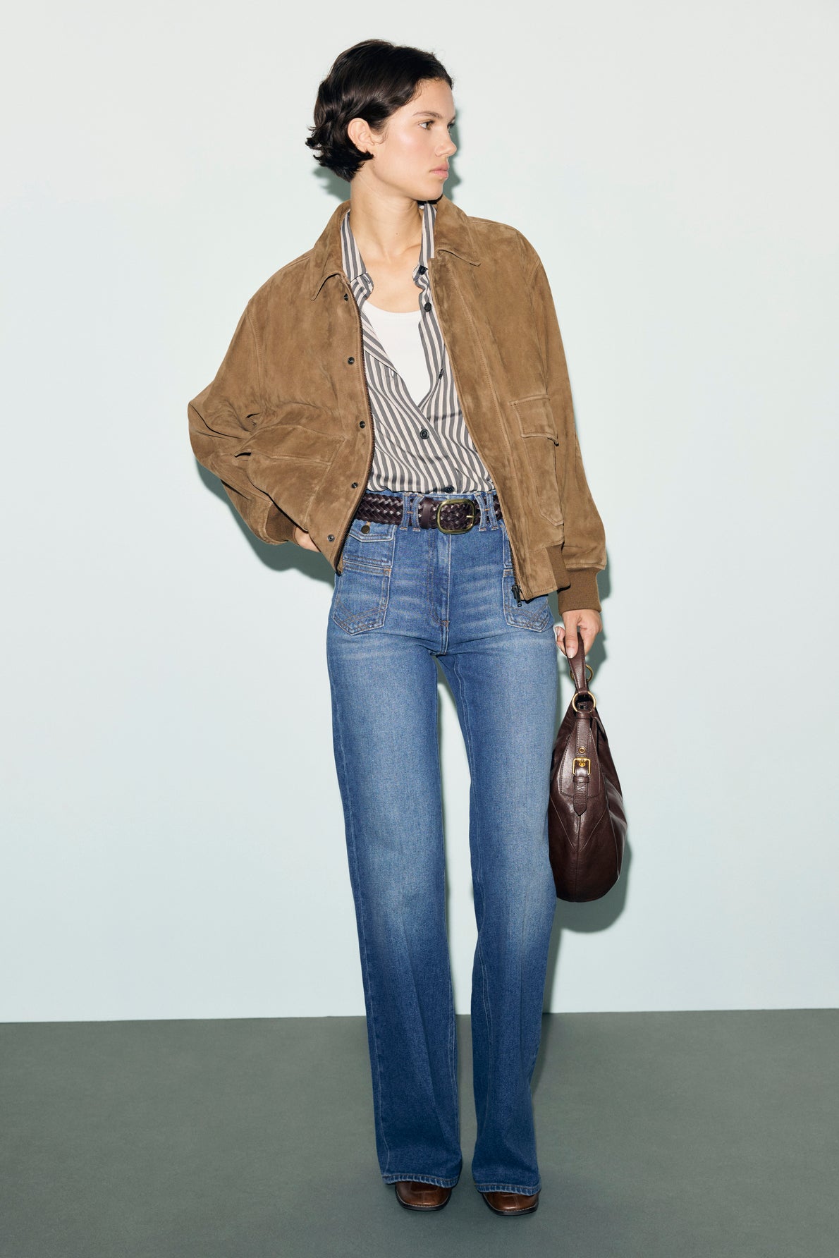 A person wearing a brown blouson cuir velours jacket, striped shirt, white tank top, blue jeans, brown belt, and brown shoes stands holding a dark brown handbag against a light background.