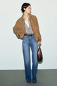 A person wearing a brown blouson cuir velours jacket, striped shirt, white tank top, blue jeans, brown belt, and brown shoes stands holding a dark brown handbag against a light background.