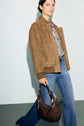 A person wearing a taupe blouson cuir velours, striped shirt, and blue jeans holds a brown leather handbag, standing against a light background.
