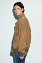 A person with short dark hair wears a blouson cuir velours in brown suede and blue jeans, holding sunglasses. They stand against a plain light background, looking over their shoulder at the camera.