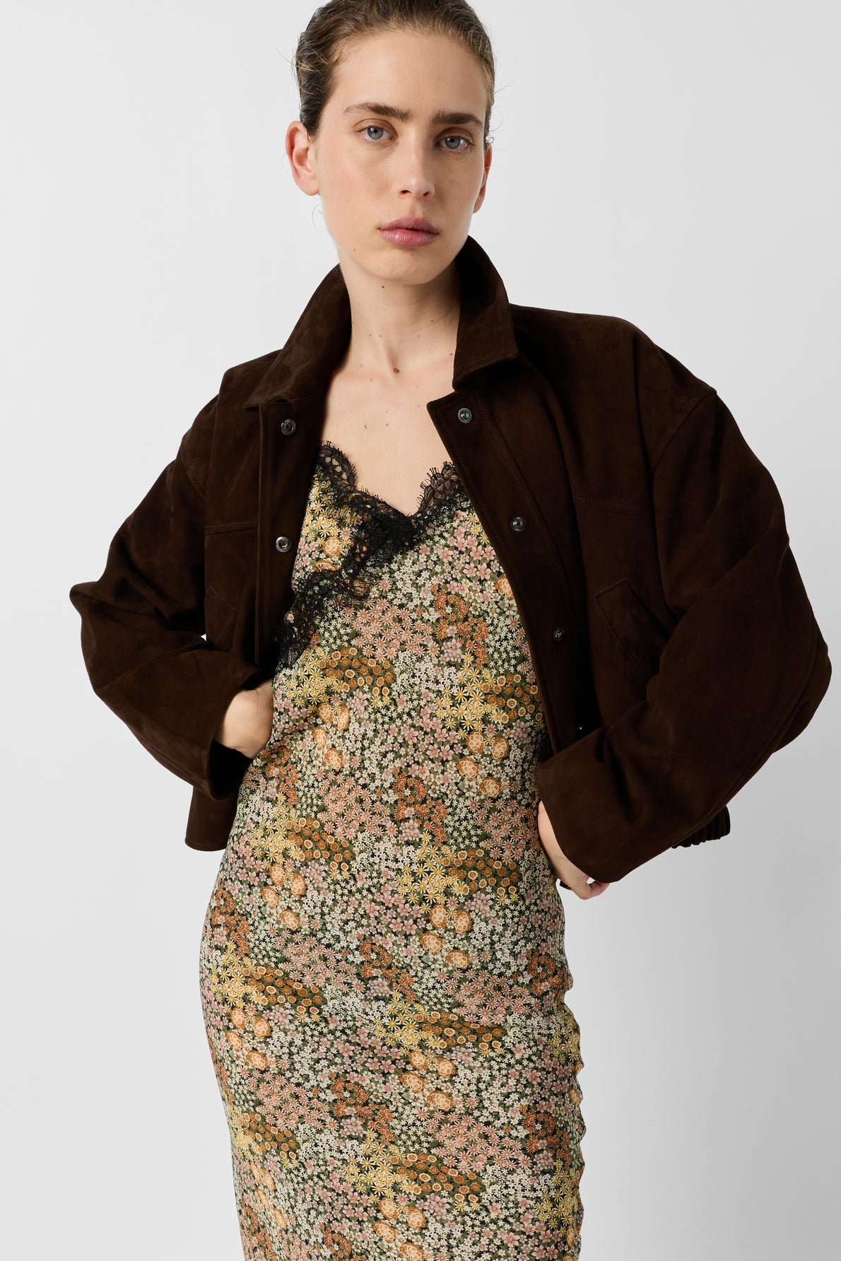 A person wearing a brown suede jacket over a floral lace-trim dress stands against a plain light background, looking confidently at the camera with hands on hips.