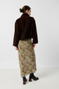 A person with brown hair in a bun wears a dark brown jacket, a long floral skirt, and black heeled boots, standing against a plain white background, facing away from the camera.