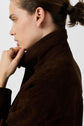 A person with light skin and brown hair in a bun wears a dark brown jacket with the collar up, holding it with one hand and looking to the side.