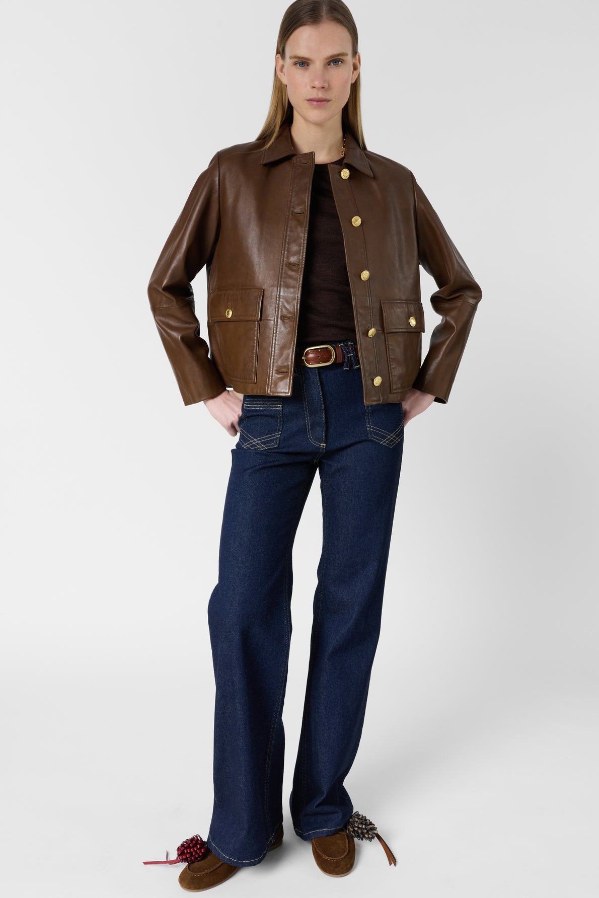 A woman stands wearing a veste en cuir vieilli, styled as a brown leather jacket, paired with a dark brown shirt, wide-leg blue jeans, and brown shoes. Her hands are tucked in her jacket pockets as she poses against a plain white background.