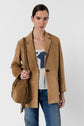 Woman wearing a tan suede blazer over a white top with blue detail, blue jeans, and carrying a tan fringed shoulder bag, standing against a plain light background.