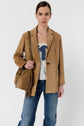 Woman with brown hair in a low bun wearing a tan suede blazer, white blouse with blue bow, blue jeans, and carrying a tan fringed shoulder bag, standing against a plain white background.