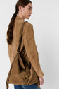 A woman with brown hair in a low ponytail wears a tan suede jacket and carries a tan suede shoulder bag with gold hardware and fringe details. She is standing against a plain white background.