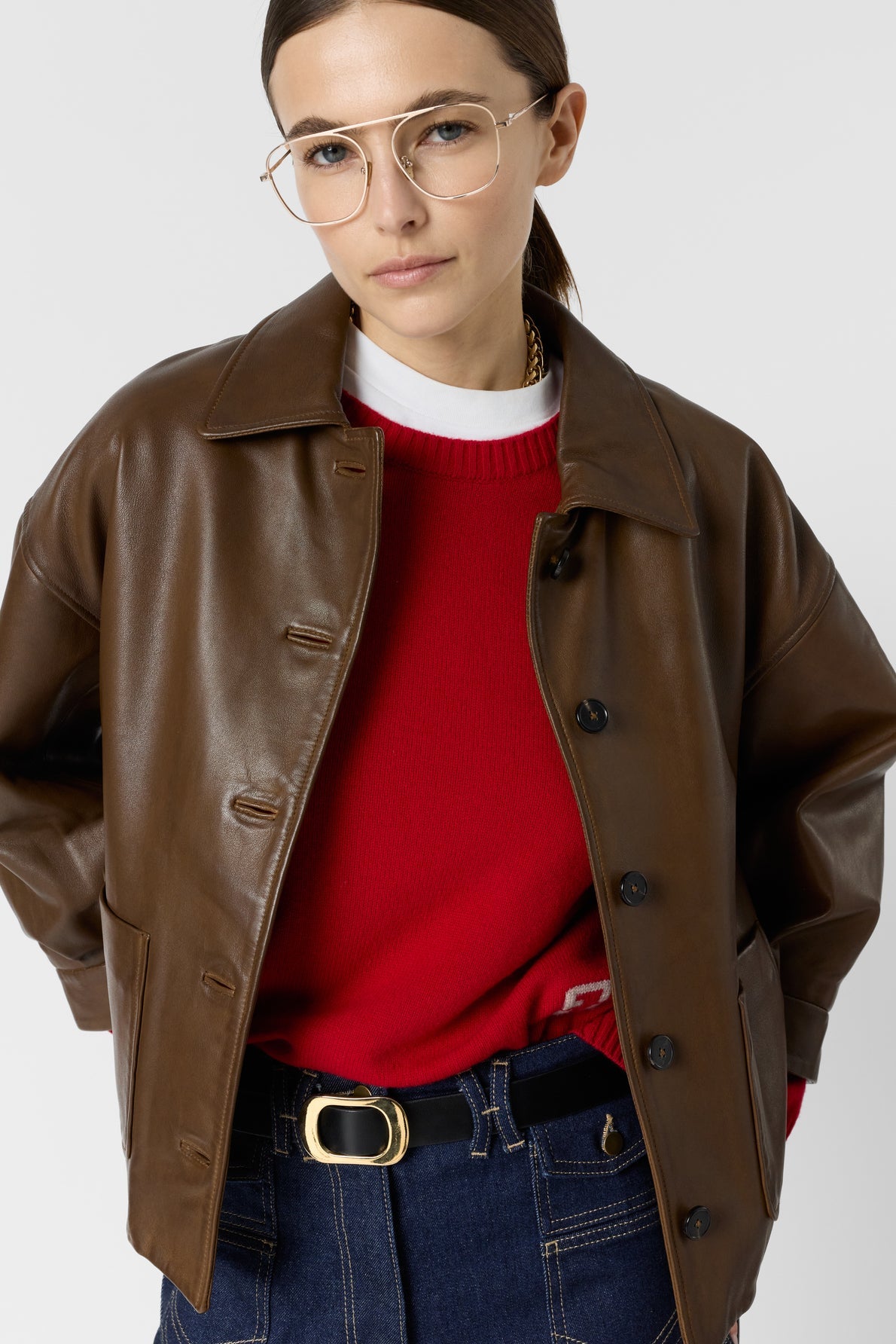 Person wearing a brown leather jacket over a red sweater, dark blue jeans with a belt, and gold-rimmed glasses, standing against a plain light background.
