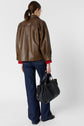A woman with a ponytail wearing a brown leather jacket, red sleeves, blue jeans, and black shoes stands facing away, holding a large black handbag in her left hand against a plain background.