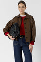 Woman wearing glasses, a brown leather jacket, red sweater, and blue jeans holds a black leather bag under her arm, standing against a plain white background.