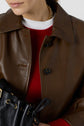 Woman wearing a brown leather jacket over a red sweater and white top, holding a black drawstring leather bag, with a gold chain necklace partially visible.