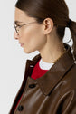 A woman with brown hair in a ponytail wears glasses, a gold chain, a brown leather jacket, and a red top with a white collar, shown in profile against a plain background.