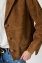 A person wearing a brown suede jacket with large buttons and a pocket, a white shirt, and blue jeans with embroidered flowers and a gold-buckled belt.