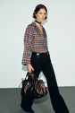 A woman with short dark hair wears a blouse Lorra à imprimé graphique, black pants, a black choker, and holds a large black handbag while walking against a plain background.