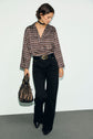 A woman wearing a blouse Lorra à imprimé graphique, black pants, and black boots holds a metallic drawstring bag. She has short hair, a black choker with col chemise detailing, and looks down with one hand in her pocket.