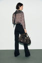 A person with short dark hair, wearing a patterned sheer long-sleeve blouse Lorra with a col chemise and black pants, stands facing away while holding a large, dark handbag. The background is plain and minimalist, highlighting the striking imprimé graphique of the blouse.