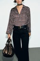 Person wearing a blouse Lorra with an imprimé graphique, black pants with a belt, and a choker, holding a metallic brown drawstring handbag, standing against a plain background.