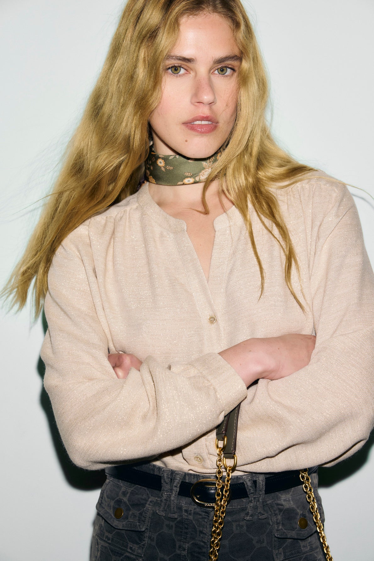 A woman with long blonde hair crosses her arms, wearing a chic light beige blouse manches longues, a patterned green scarf, and dark pants with a gold chain purse. She stands against a plain white background.