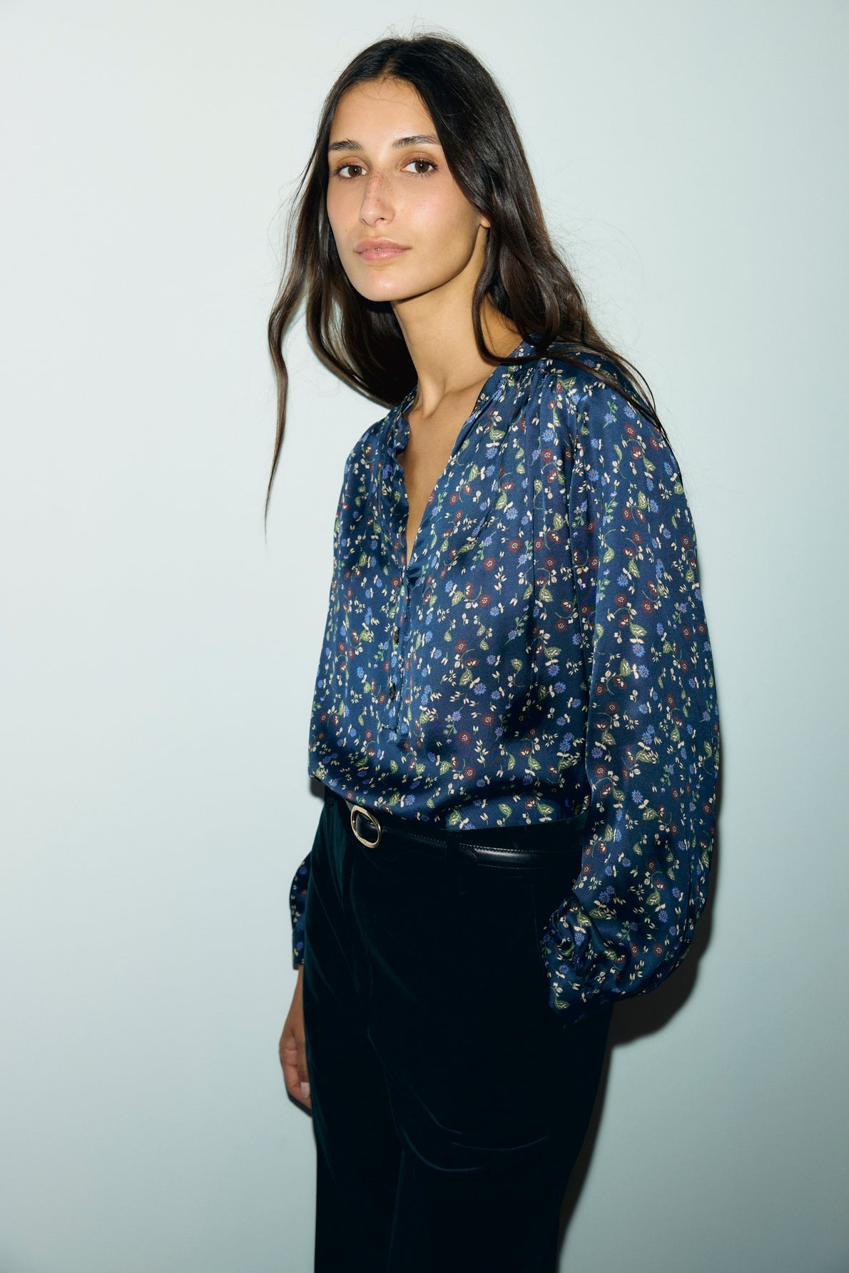 A woman with long dark hair wears a blue motif floral blouse Lolita with puffed manches longues and black pants, standing against a plain light background.
