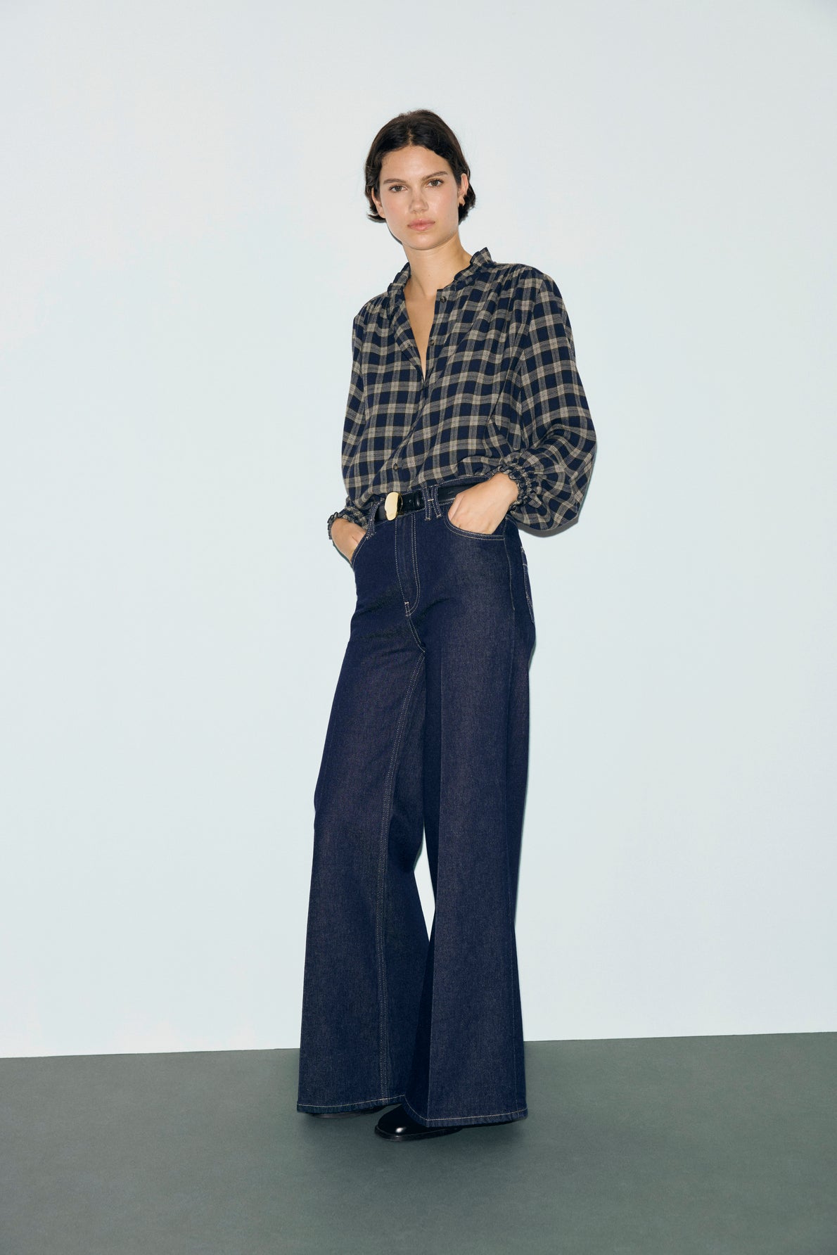 A person stands with hands in pockets, wearing a chemise à carreaux in black and gray tucked into high-waisted, wide-leg dark denim jeans, against a plain background.
