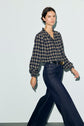 A woman walks wearing a navy chemise à carreaux with a col montant à volants and poignets smockés, tucked into high-waisted dark wide-leg jeans with a round belt buckle, against a light background.