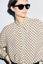 A close-up studio shot of a woman with dark hair in a bun wearing black, thick-framed sunglasses and a beige chemise fluide featuring a repeating black geometric interlocking logo pattern. The motif imprimé adds a sophisticated touch to her contemporary look.