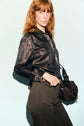 A woman with red hair wears a shiny, dark chemise satinée and high-waisted dark pants, holding a small black handbag. She stands against a light background, looking seriously at the camera.