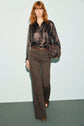A woman with wavy red hair wears a chemise satinée noire, brown belted pants, and brown shoes. She holds a brown handbag and stands against a pale blue background.