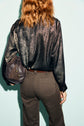 A person with wavy, shoulder-length red hair is seen from behind, wearing a chemise satinée noire and brown pants. They hold a small brown leather bag in their left hand against a light blue background.