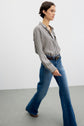 A person walks indoors wearing a chemise à rayures black-and-white tucked into high-waisted blue jeans with a braided belt. The background is plain and minimalistic.